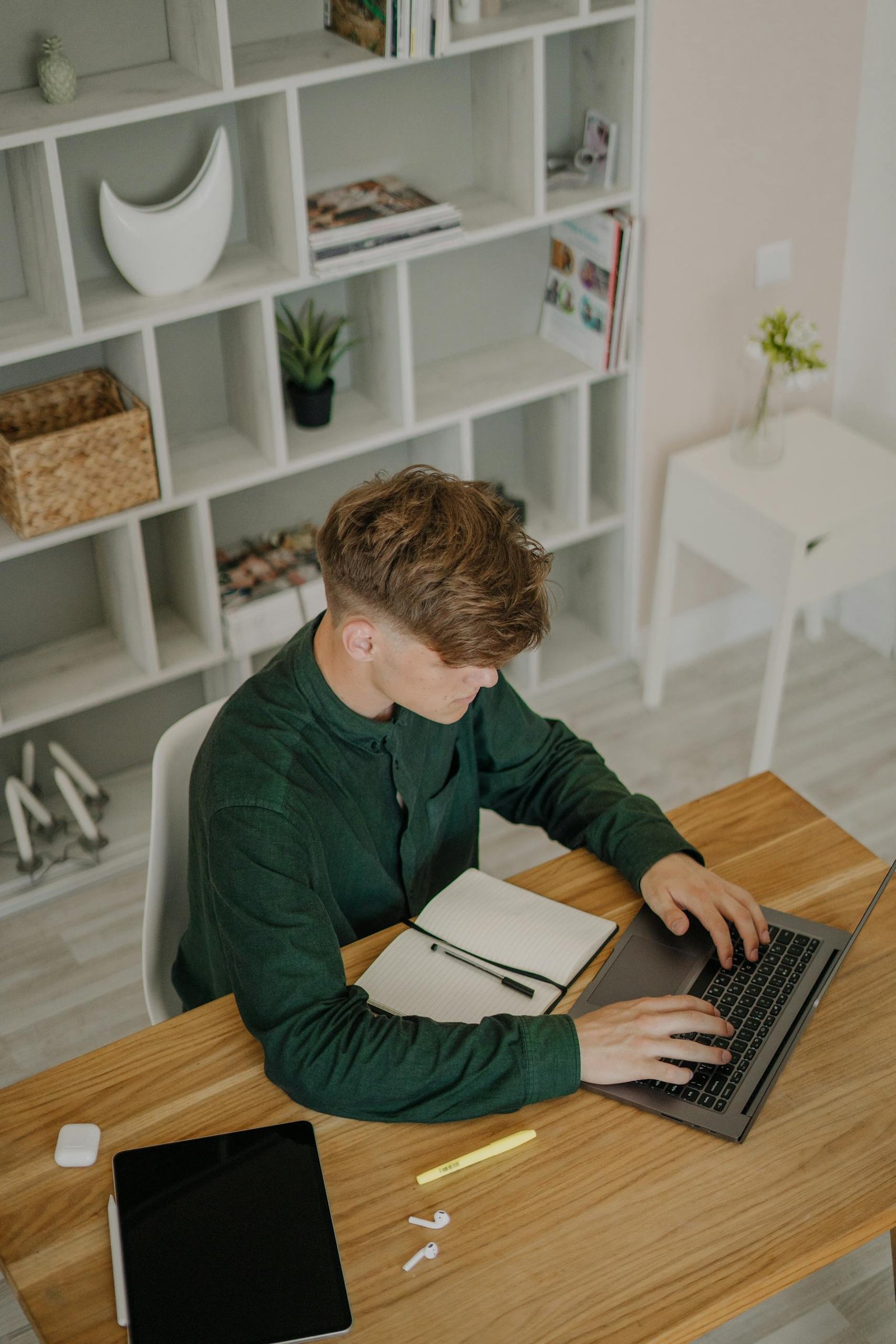 High-Angle Shot of a Male Student in Green Long Sleeves Using a Laptop on a Wooden Desk